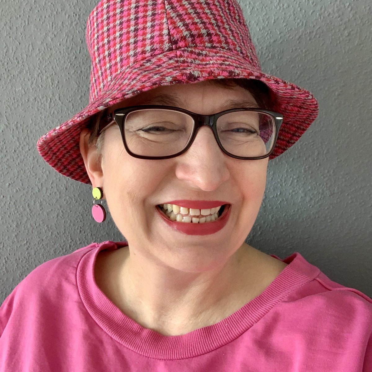Iris lachend mit pinkem Hut und zweifarbigen Ohrhängern in Neongelb und Pink