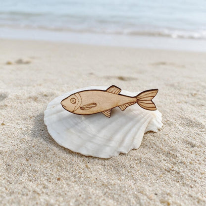 Sprotten-Brosche aus Holz in Natur auf einer Muschel am Strand